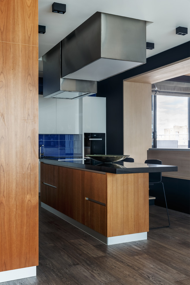 Expansive contemporary l-shaped open plan kitchen in Moscow with an undermount sink, flat-panel cabinets, white cabinets, granite benchtops, blue splashback, glass tile splashback, black appliances, medium hardwood floors, with island, brown floor and black benchtop.