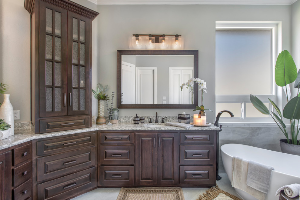This is an example of a mid-sized traditional master bathroom in Other with raised-panel cabinets, brown cabinets, a freestanding tub, a corner shower, a two-piece toilet, gray tile, ceramic tile, grey walls, ceramic floors, an undermount sink, quartzite benchtops, grey floor, a hinged shower door, grey benchtops, an enclosed toilet, a double vanity and a built-in vanity.