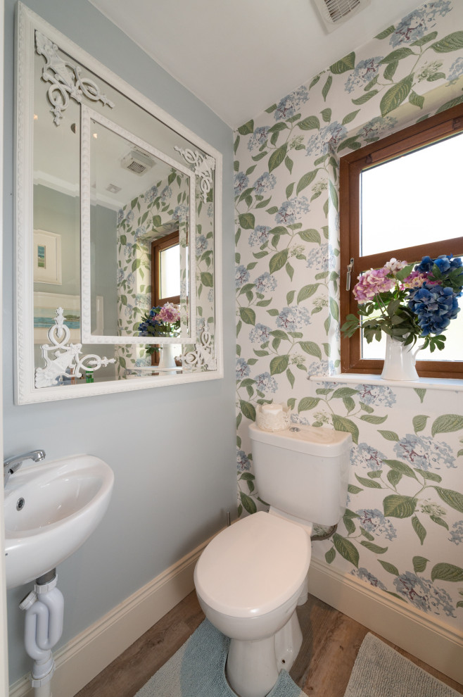 Photo of a transitional powder room in Cork with open cabinets, white cabinets, a two-piece toilet, grey walls, medium hardwood floors, a wall-mount sink, brown floor, white benchtops, a floating vanity and wallpaper.