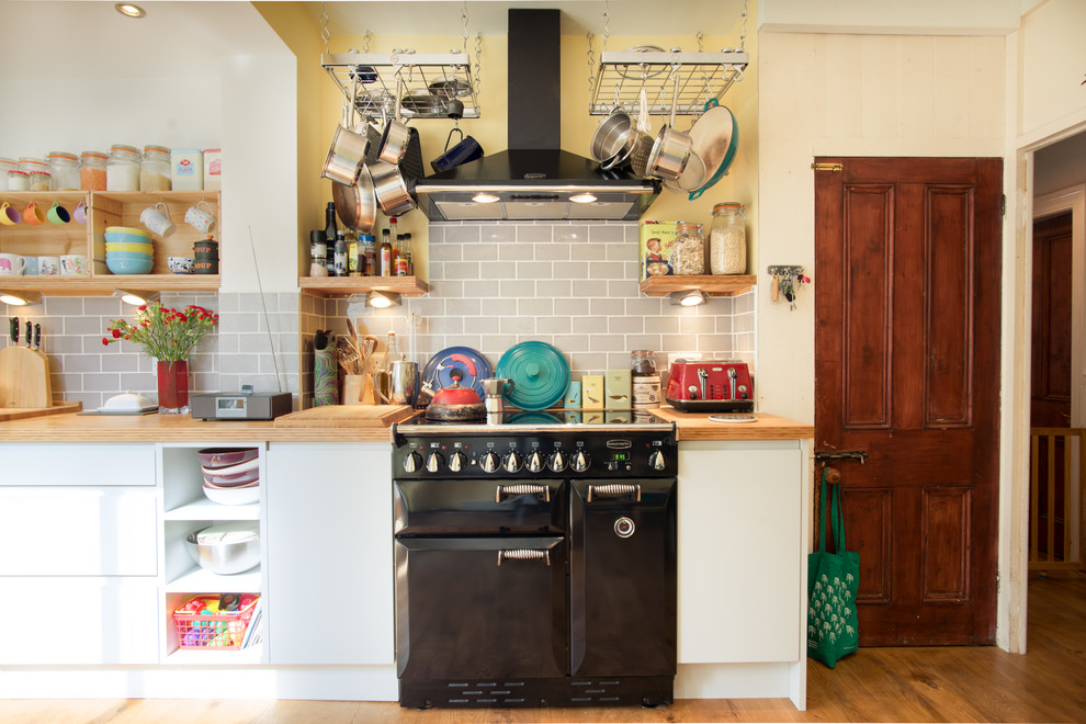 Inspiration for a mid-sized eclectic l-shaped eat-in kitchen in Other with a farmhouse sink, flat-panel cabinets, grey cabinets, wood benchtops, grey splashback, subway tile splashback, black appliances, medium hardwood floors and no island.