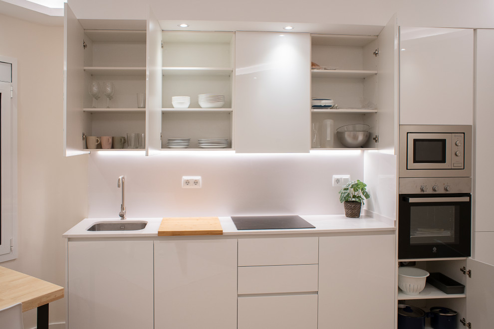 Photo of a mid-sized contemporary single-wall open plan kitchen in Barcelona with a single-bowl sink, flat-panel cabinets, white cabinets, white splashback, stainless steel appliances, medium hardwood floors, no island, brown floor and white benchtop.