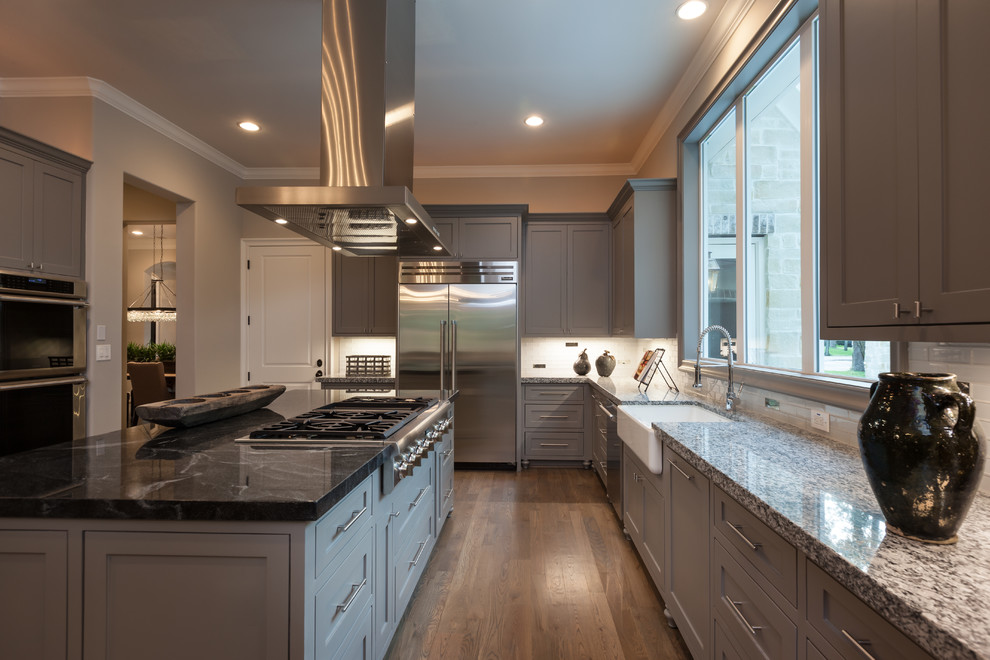 Design ideas for a large transitional u-shaped open plan kitchen in Houston with a farmhouse sink, shaker cabinets, grey cabinets, granite benchtops, white splashback, glass tile splashback, stainless steel appliances, light hardwood floors and with island.