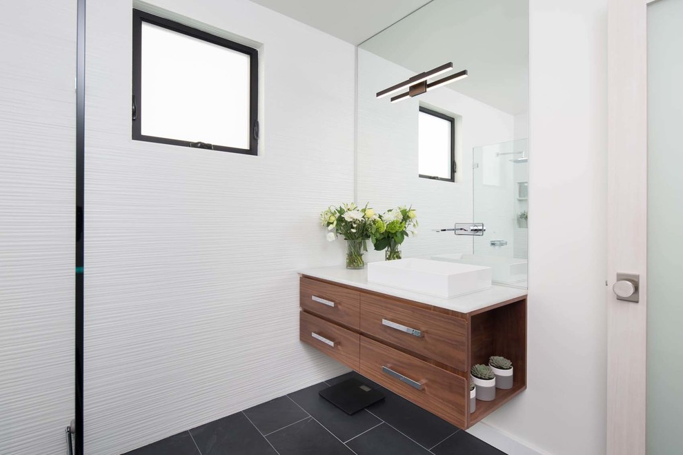 Photo of a mid-sized contemporary kids bathroom in Los Angeles with flat-panel cabinets, brown cabinets, a curbless shower, a wall-mount toilet, gray tile, stone tile, white walls, slate floors, a vessel sink, glass benchtops, black floor, an open shower and white benchtops.
