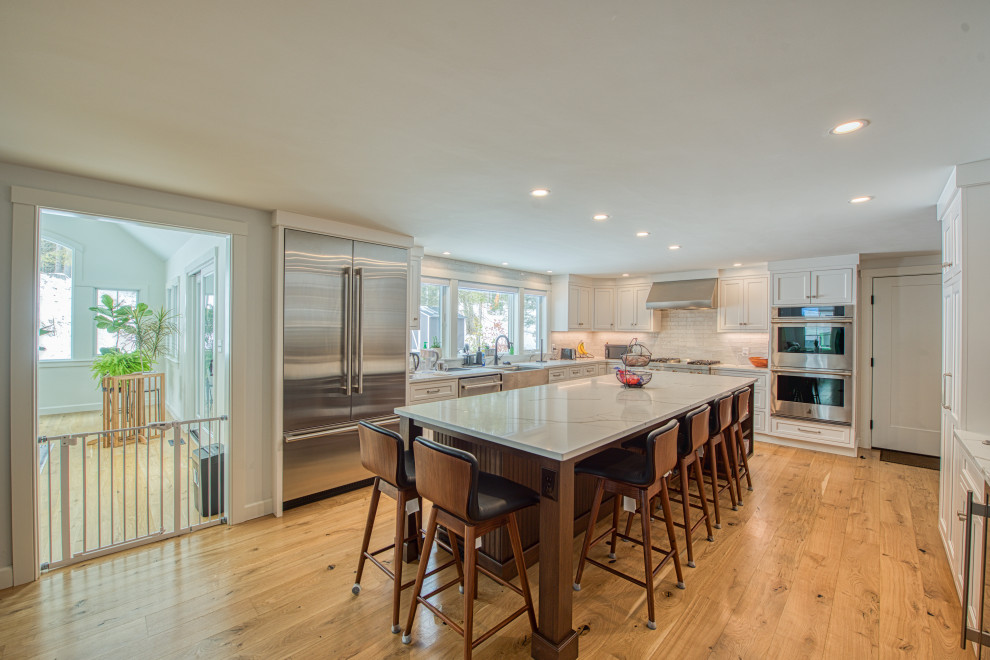 Transitional light wood floor kitchen photo with a farmhouse sink, shaker cabinets, white cabinets, quartz countertops, white backsplash, ceramic backsplash, stainless steel appliances, an island and white countertops