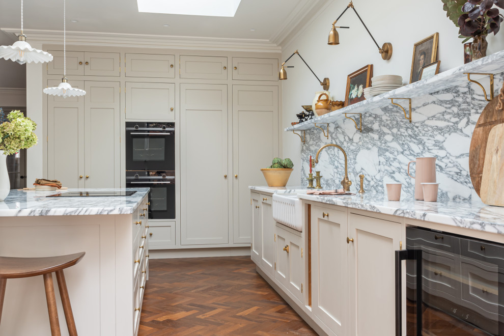 Mid-sized traditional eat-in kitchen in West Midlands with a farmhouse sink, shaker cabinets, beige cabinets, marble benchtops, white splashback, marble splashback, black appliances, medium hardwood floors, with island, brown floor and white benchtop.