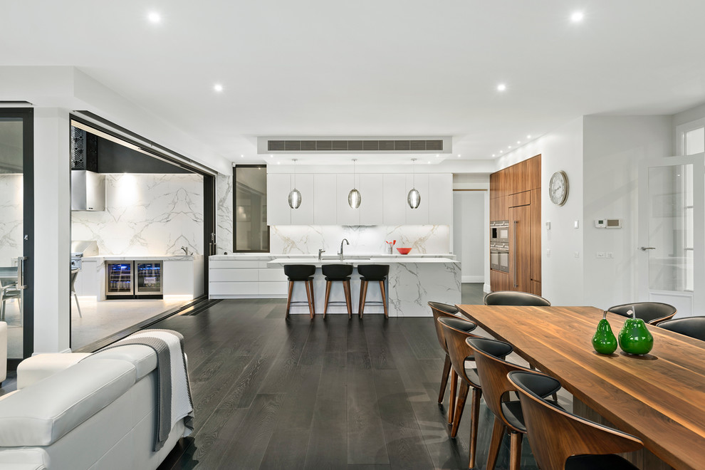 Photo of a large contemporary galley open plan kitchen in Melbourne with an integrated sink, flat-panel cabinets, white cabinets, marble benchtops, white splashback, stone slab splashback, black appliances, dark hardwood floors and with island.