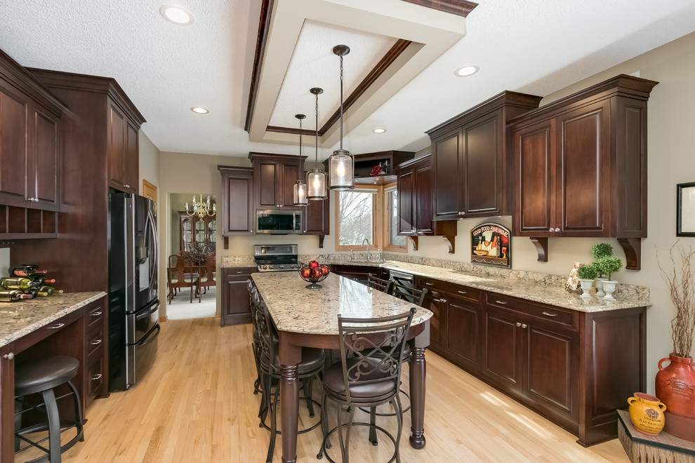 This is an example of a large traditional u-shaped eat-in kitchen in Minneapolis with an undermount sink, raised-panel cabinets, dark wood cabinets, granite benchtops, stainless steel appliances, light hardwood floors, with island, beige floor and beige benchtop.
