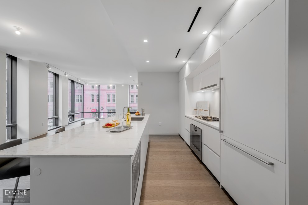 Photo of a large contemporary single-wall open plan kitchen in Boston with an undermount sink, flat-panel cabinets, white cabinets, quartz benchtops, white splashback, glass sheet splashback, panelled appliances, light hardwood floors, with island and beige floor.