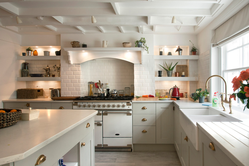 This is an example of a large traditional l-shaped kitchen in London with a farmhouse sink, shaker cabinets, grey cabinets, granite benchtops, white splashback, subway tile splashback, with island, white benchtop, white appliances and light hardwood floors.