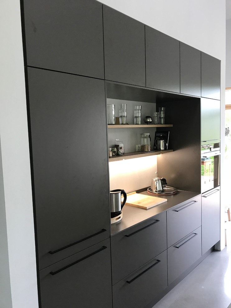 Geräumige, Zweizeilige Industrial Wohnküche mit integriertem Waschbecken, flächenbündigen Schrankfronten, grauen Schränken, Betonarbeitsplatte, Küchenrückwand in Weiß, schwarzen Elektrogeräten, Betonboden, Kücheninsel, grauem Boden und grauer Arbeitsplatte in Sonstige