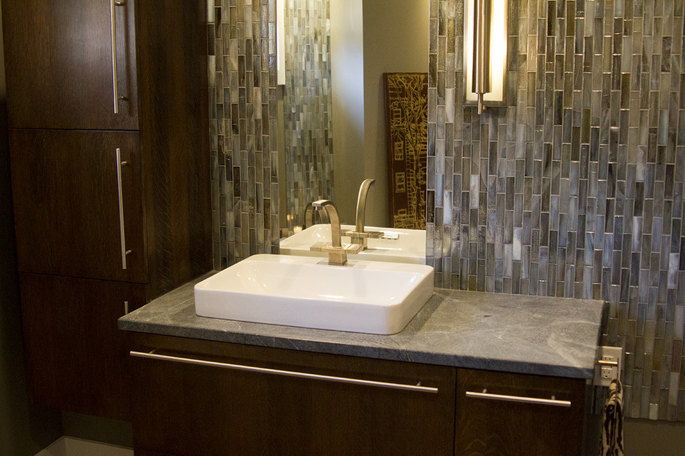 Photo of a mid-sized midcentury master bathroom in Omaha with flat-panel cabinets, dark wood cabinets, a freestanding tub, blue tile, brown tile, multi-coloured tile, mosaic tile, brown walls, porcelain floors, a vessel sink, soapstone benchtops and beige floor.