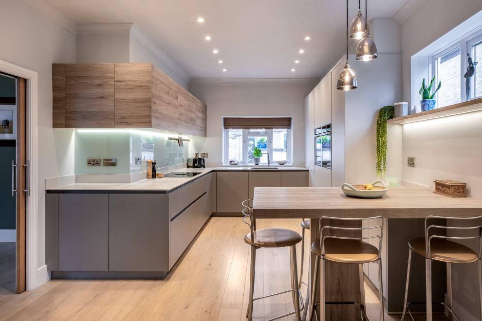 Design ideas for a medium sized contemporary u-shaped kitchen/diner in Gloucestershire with a built-in sink, flat-panel cabinets, blue splashback, glass sheet splashback, black appliances, light hardwood flooring and brown floors.