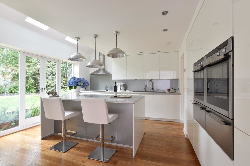 Design ideas for a mid-sized contemporary galley kitchen in Manchester with a double-bowl sink, flat-panel cabinets, white cabinets, quartz benchtops, grey splashback, glass sheet splashback, stainless steel appliances, medium hardwood floors and with island.