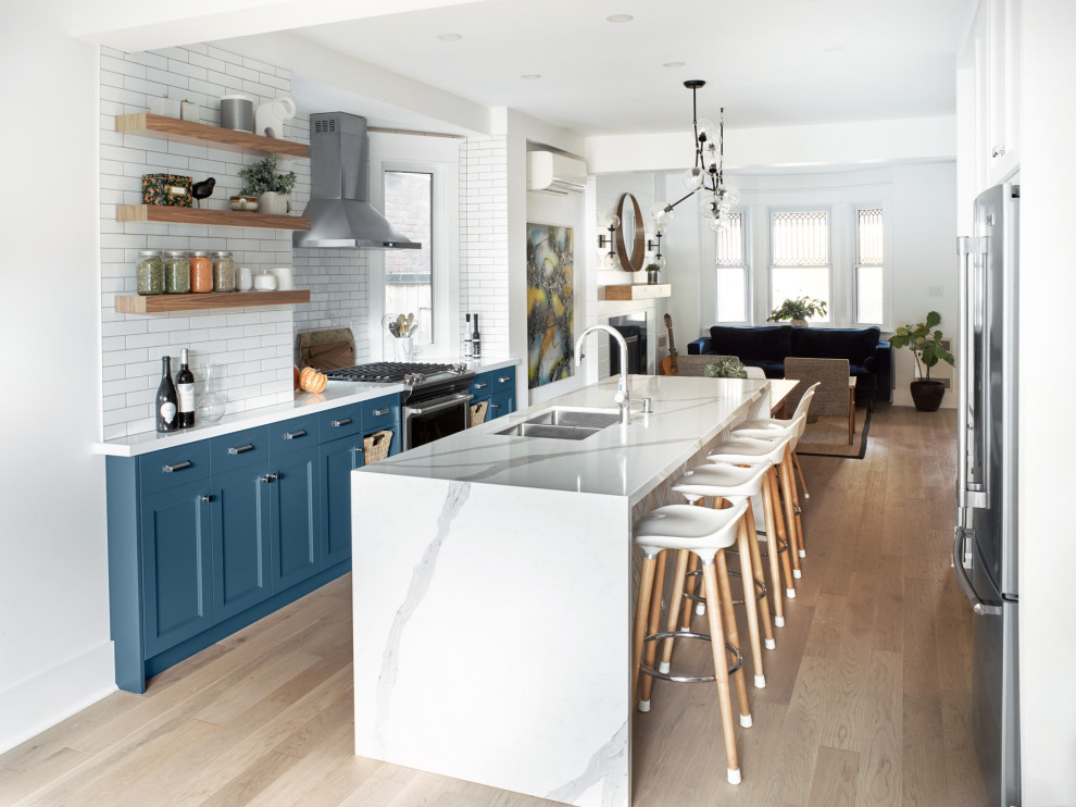 This is an example of a large transitional galley open plan kitchen in Toronto with a double-bowl sink, shaker cabinets, blue cabinets, quartz benchtops, white splashback, subway tile splashback, stainless steel appliances, light hardwood floors, with island, brown floor and white benchtop.