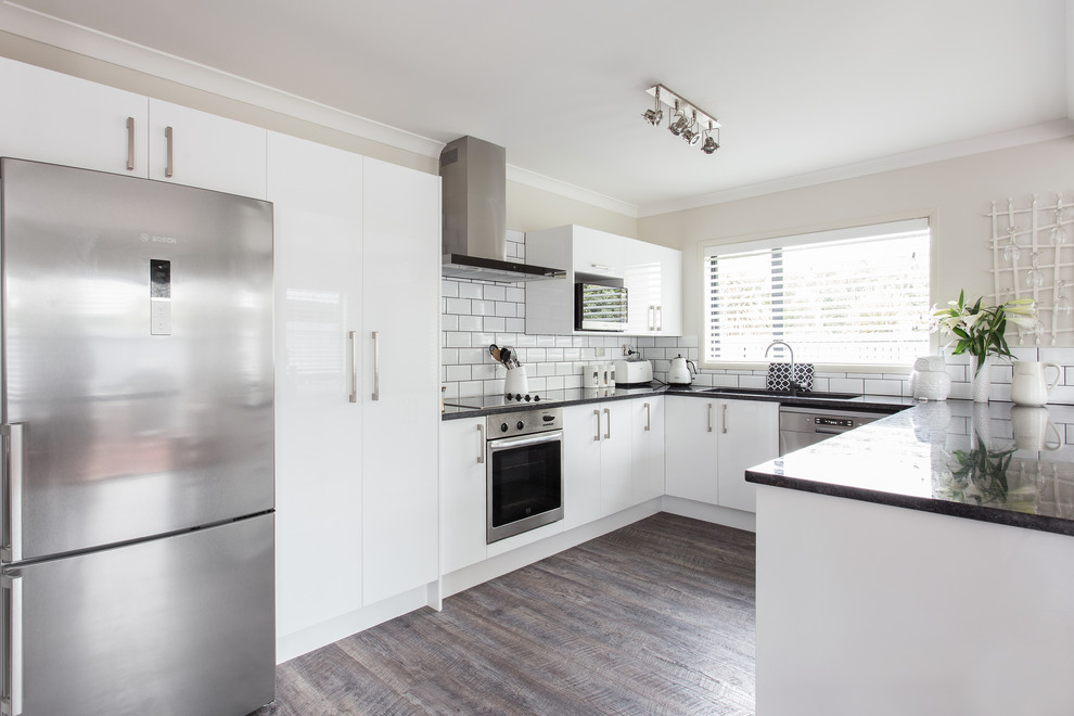 This is an example of a mid-sized modern u-shaped eat-in kitchen in Christchurch with an undermount sink, flat-panel cabinets, white cabinets, granite benchtops, white splashback, ceramic splashback, stainless steel appliances, laminate floors, a peninsula, brown floor and black benchtop.