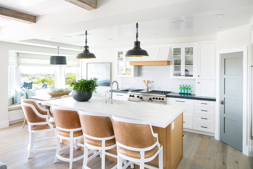 Design ideas for a beach style galley eat-in kitchen in Orange County with a farmhouse sink, shaker cabinets, white cabinets, white splashback, subway tile splashback, stainless steel appliances, dark hardwood floors, with island, brown floor and black benchtop.