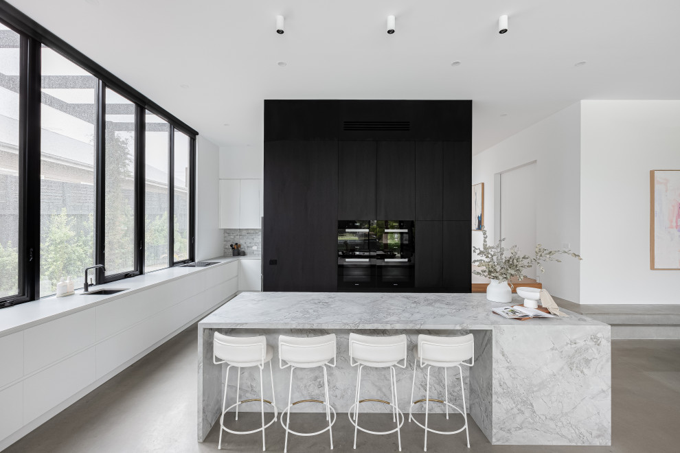 Cette image montre une cuisine ouverte minimaliste en L avec un évier encastré, un placard à porte plane, des portes de placard blanches, une crédence grise, un électroménager noir, sol en béton ciré, îlot, un sol gris et un plan de travail gris.