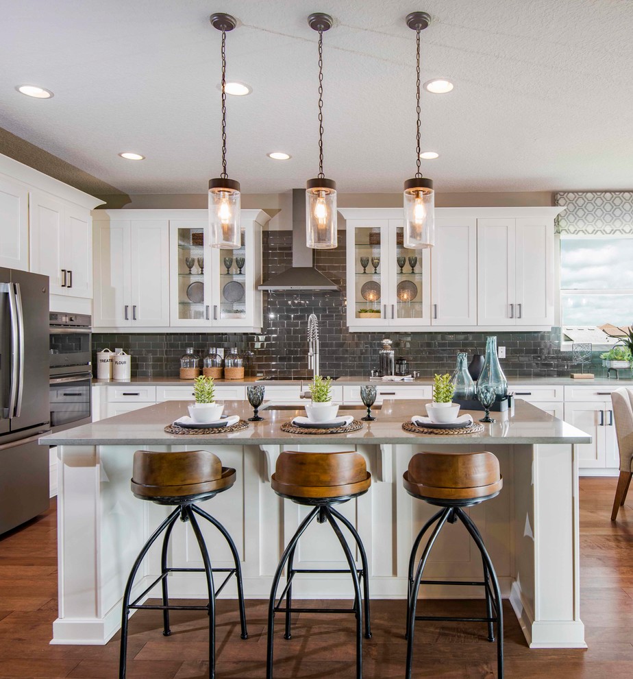 Photo of a country l-shaped eat-in kitchen in Orlando with white cabinets, quartzite benchtops, grey splashback, glass tile splashback, stainless steel appliances, medium hardwood floors, with island, brown floor, grey benchtop and shaker cabinets.