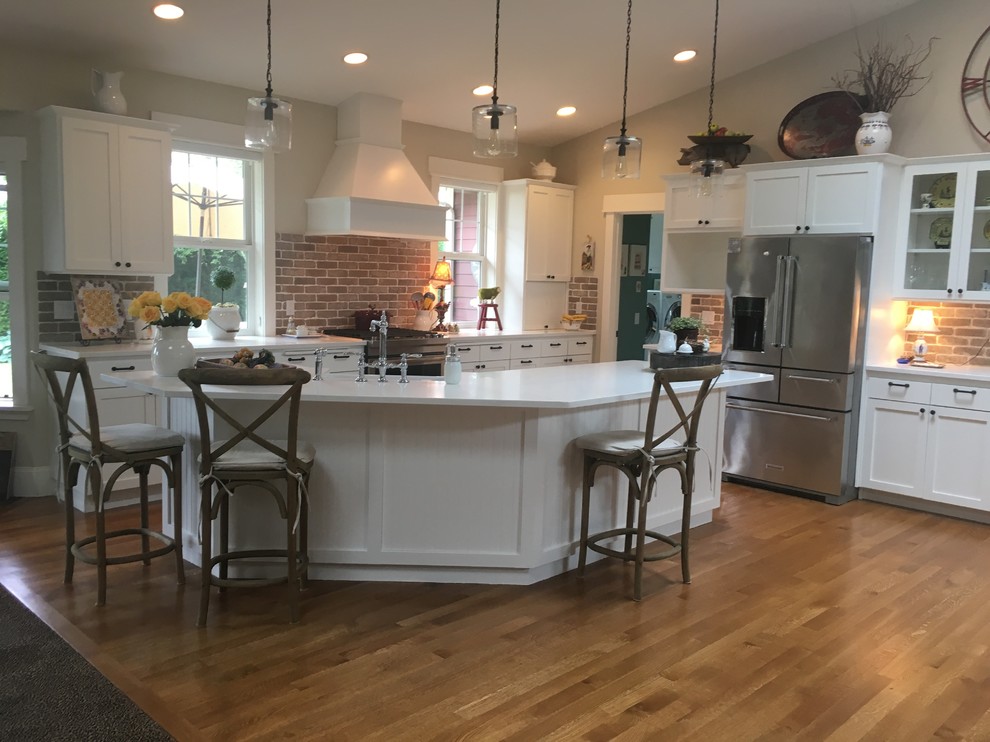 Photo of a large country kitchen with a farmhouse sink, shaker cabinets, white cabinets, quartz benchtops, red splashback, brick splashback, stainless steel appliances, light hardwood floors, with island and white benchtop.