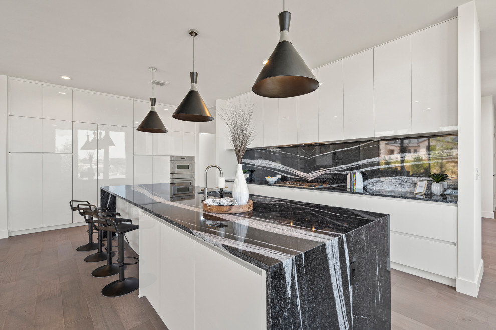 Photo of a mid-sized contemporary galley eat-in kitchen in Austin with an undermount sink, flat-panel cabinets, white cabinets, granite benchtops, black splashback, granite splashback, stainless steel appliances, medium hardwood floors, with island, brown floor and black benchtop.