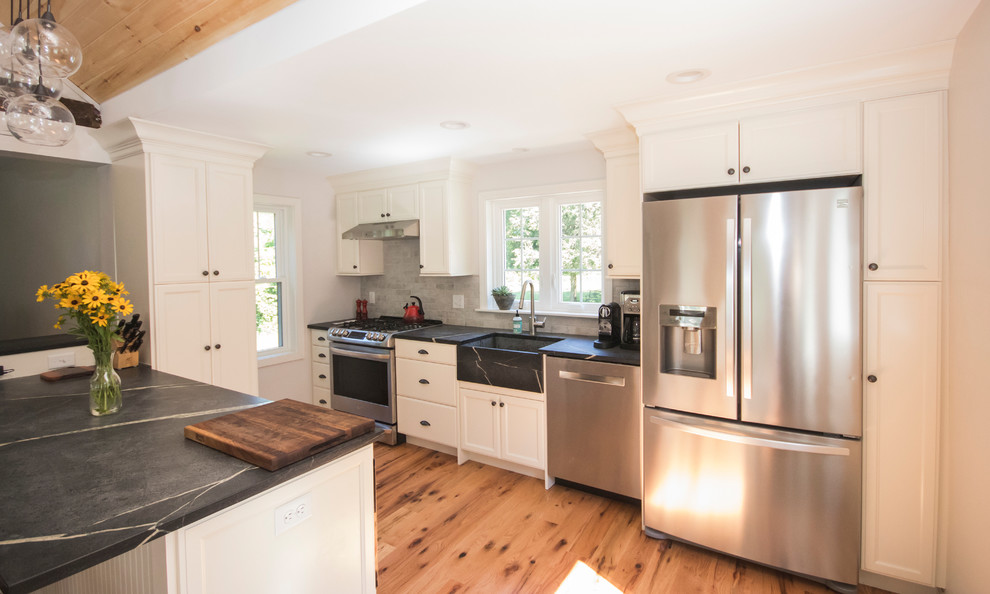 Design ideas for an expansive country l-shaped eat-in kitchen in Providence with a farmhouse sink, recessed-panel cabinets, white cabinets, soapstone benchtops, white splashback, porcelain splashback, stainless steel appliances, light hardwood floors, with island and beige floor.