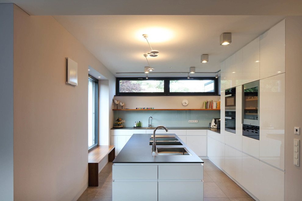 This is an example of a mid-sized contemporary l-shaped open plan kitchen in Stuttgart with with island, a drop-in sink, flat-panel cabinets, white cabinets, blue splashback, glass sheet splashback, black appliances, cement tiles and beige floor.