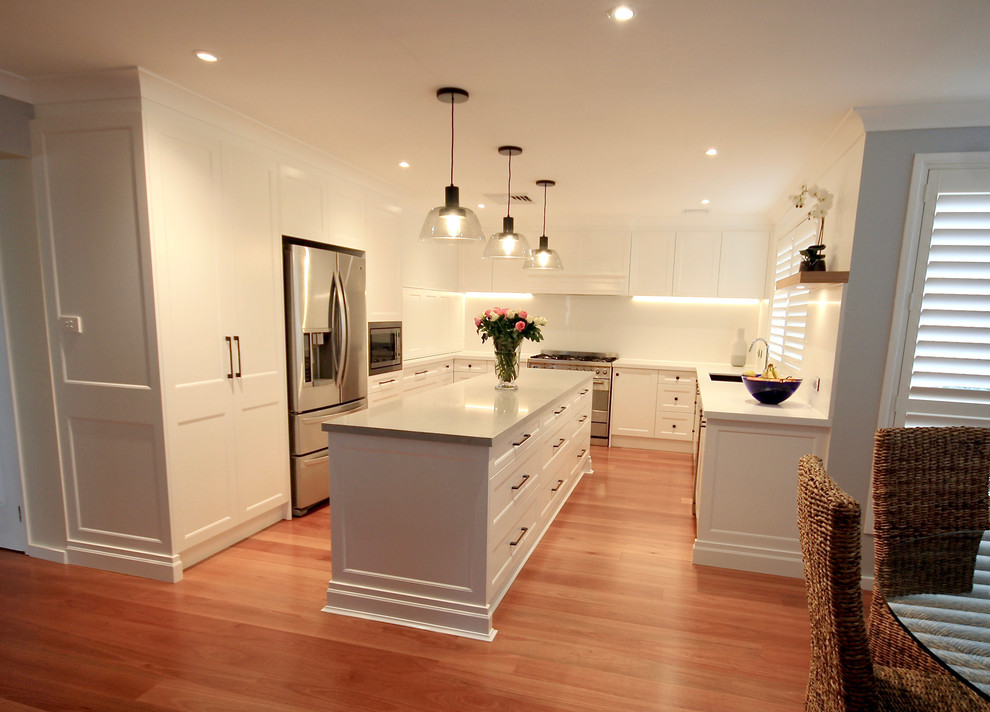 This is an example of a large transitional u-shaped eat-in kitchen in Sydney with an undermount sink, recessed-panel cabinets, white cabinets, quartz benchtops, white splashback, stone slab splashback, stainless steel appliances, medium hardwood floors, with island, brown floor and white benchtop.