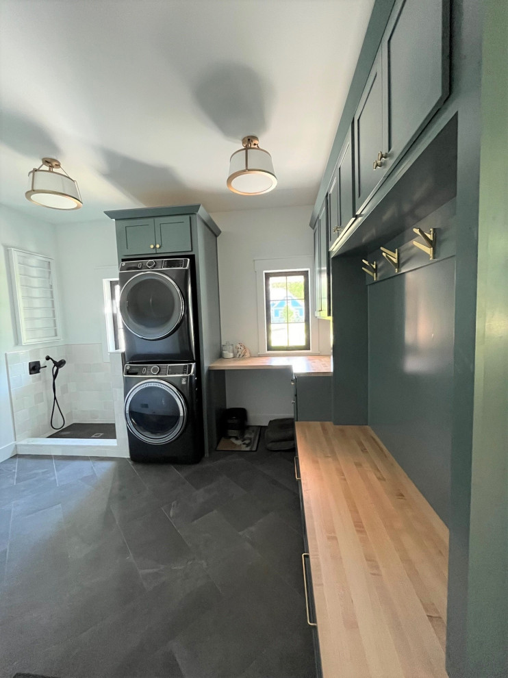 Large transitional l-shaped utility room in Detroit with shaker cabinets, green cabinets, wood benchtops, white splashback, ceramic splashback, white walls, porcelain floors, a stacked washer and dryer, grey floor and beige benchtop.