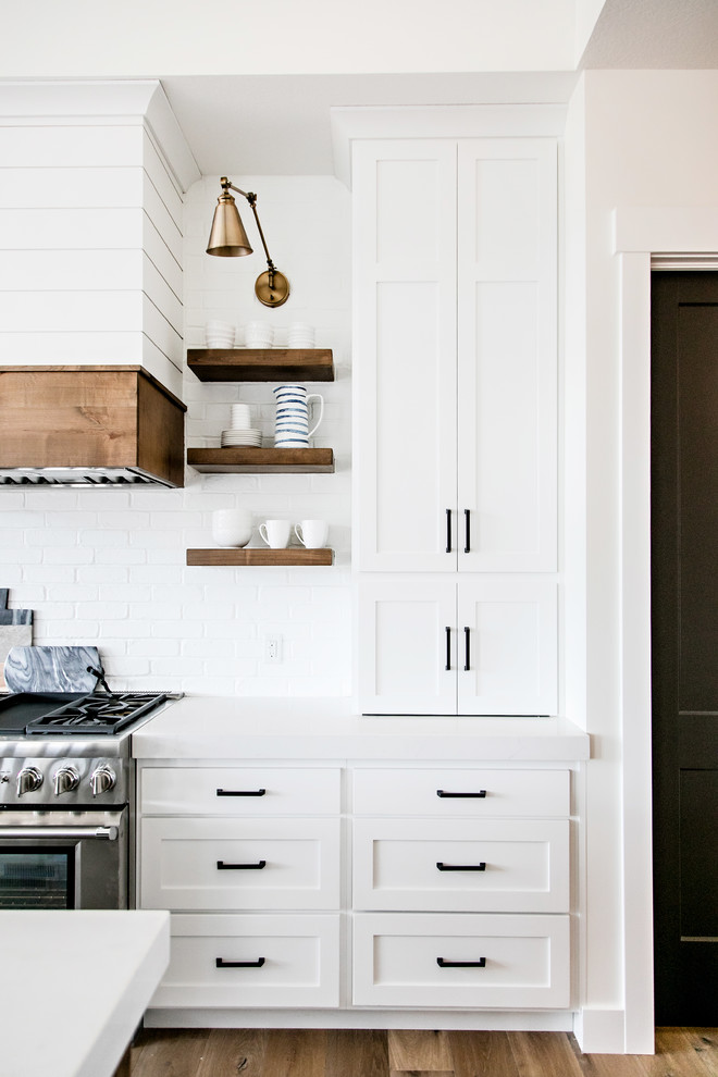 Mid-sized country kitchen in Salt Lake City with a farmhouse sink, shaker cabinets, white cabinets, quartz benchtops, white splashback, subway tile splashback, stainless steel appliances, medium hardwood floors, with island, brown floor and white benchtop.