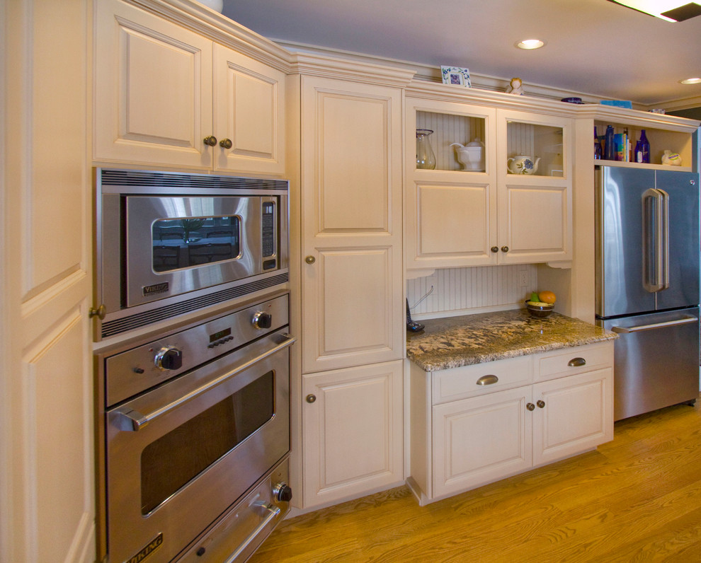 Photo of a mid-sized transitional galley separate kitchen in Other with a farmhouse sink, raised-panel cabinets, white cabinets, granite benchtops, beige splashback, stone tile splashback, stainless steel appliances, light hardwood floors and no island.