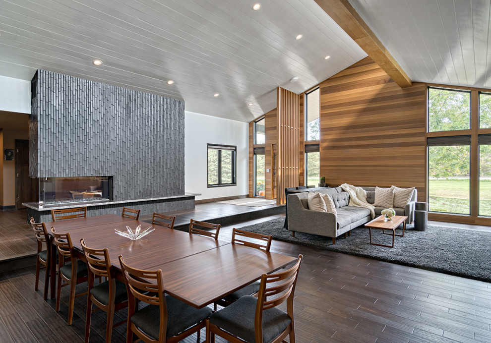 Expansive midcentury open concept living room in Minneapolis with a library, white walls, medium hardwood floors, a two-sided fireplace, a tile fireplace surround, a wall-mounted tv, brown floor and vaulted.