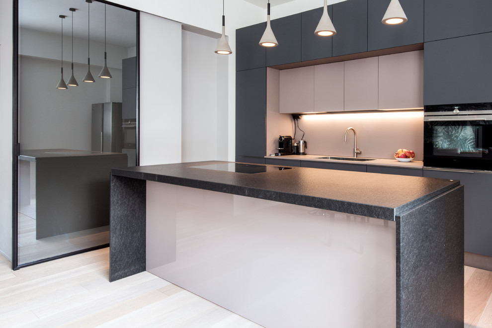 Inspiration for a large contemporary galley open plan kitchen in Milan with flat-panel cabinets, black appliances, light hardwood floors, with island, black benchtop, a drop-in sink, grey cabinets, marble benchtops, pink splashback, glass sheet splashback and beige floor.
