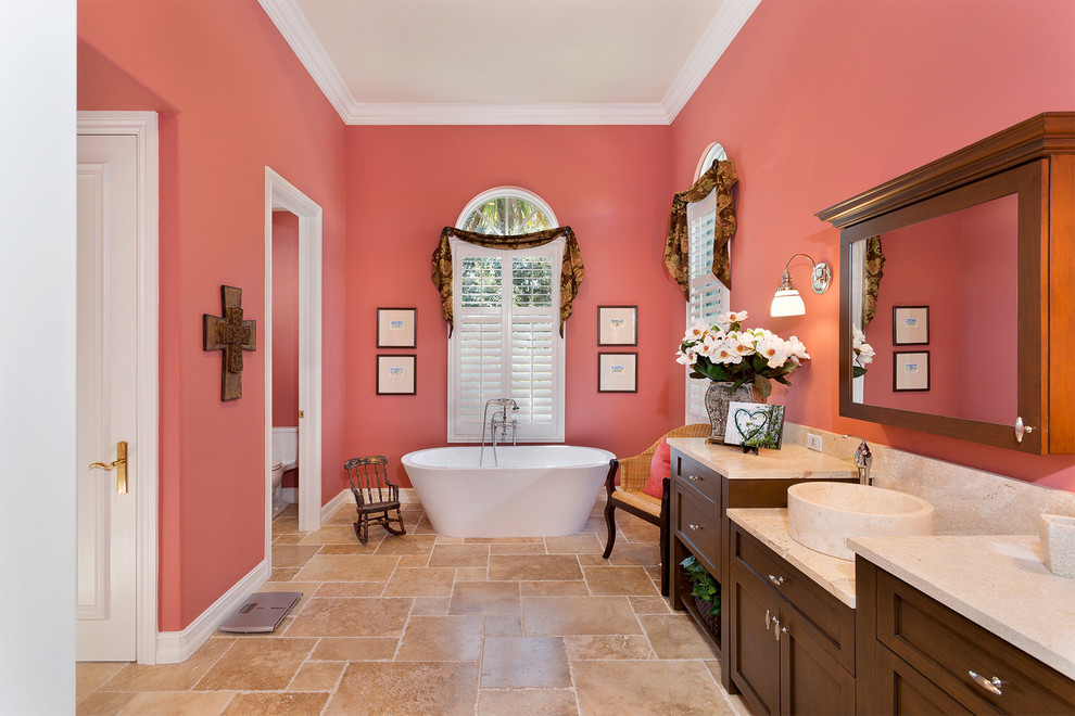 Photo of a mid-sized mediterranean master bathroom in Miami with recessed-panel cabinets, dark wood cabinets, a freestanding tub, a one-piece toilet, pink walls, travertine floors, a vessel sink, marble benchtops, beige floor and beige benchtops.