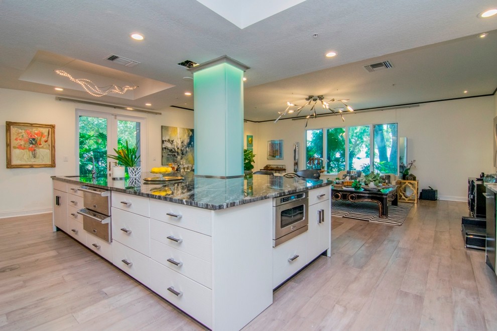 Expansive contemporary l-shaped open plan kitchen in Tampa with a double-bowl sink, flat-panel cabinets, grey cabinets, granite benchtops, white splashback, stone slab splashback, black appliances, light hardwood floors and with island.