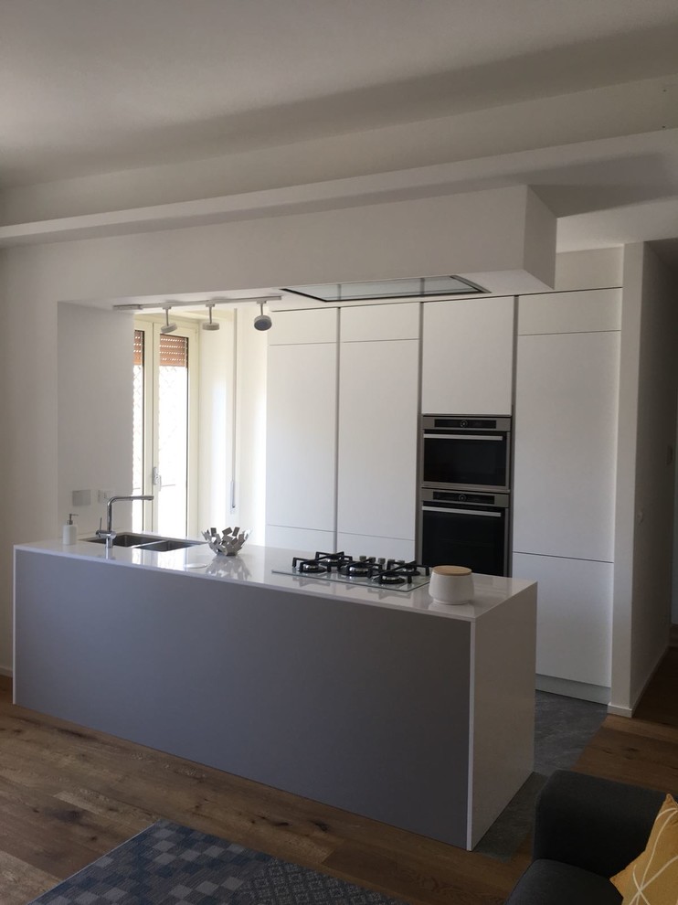 Photo of a mid-sized modern galley open plan kitchen in Rome with an undermount sink, flat-panel cabinets, grey cabinets, quartz benchtops, white appliances, light hardwood floors, with island, brown floor and white benchtop.