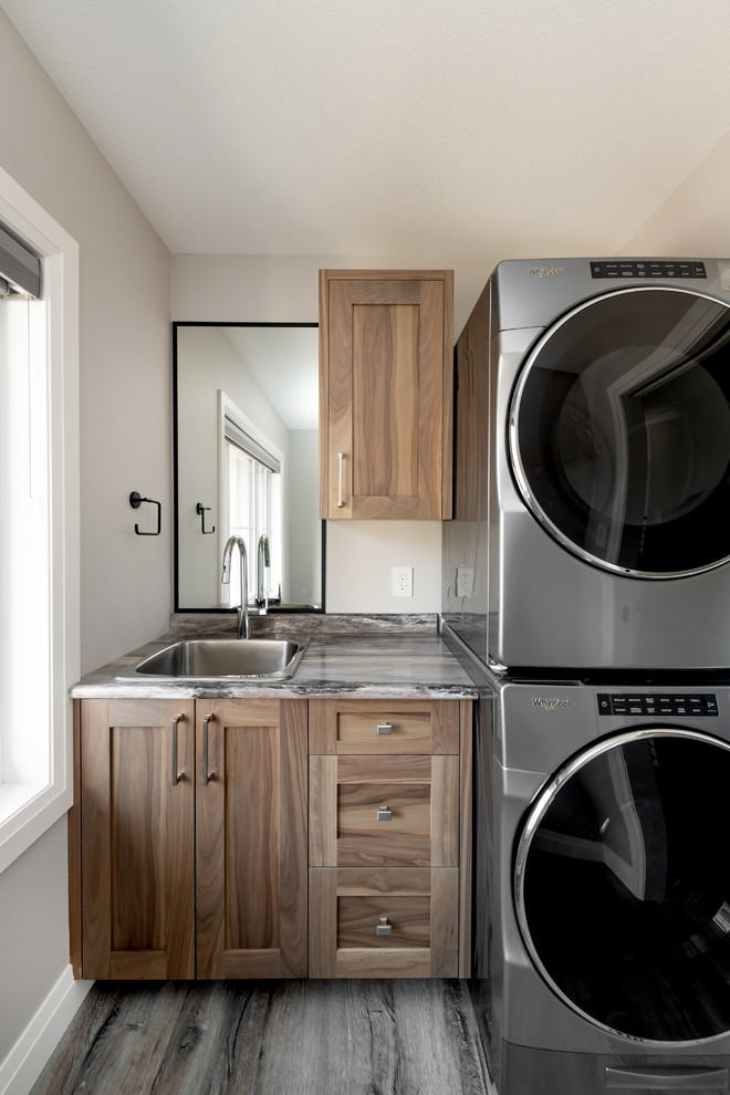 This is an example of a mid-sized transitional single-wall utility room in Other with a single-bowl sink, shaker cabinets, medium wood cabinets, laminate benchtops, grey walls, vinyl floors, a stacked washer and dryer, brown floor and multi-coloured benchtop.