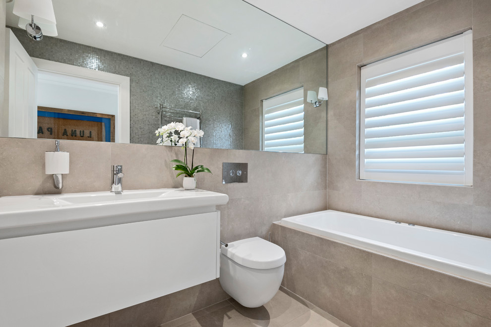 This is an example of a large modern master bathroom in Sydney with flat-panel cabinets, white cabinets, a wall-mount toilet, beige tile, stone tile, beige walls, porcelain floors, a console sink, solid surface benchtops, beige floor and white benchtops.