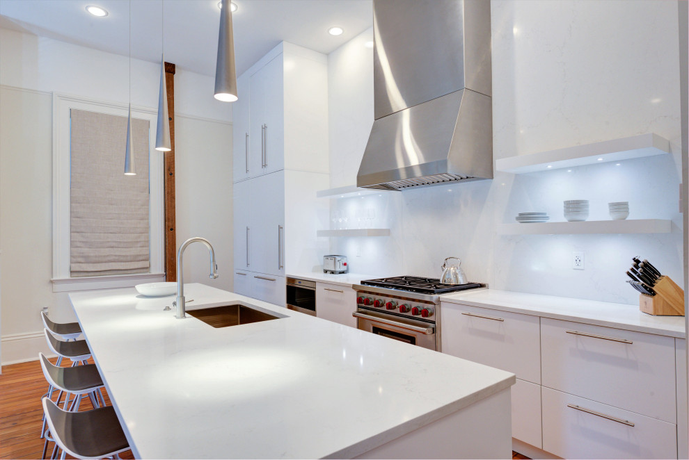 Photo of a mid-sized contemporary galley open plan kitchen in New Orleans with flat-panel cabinets, white cabinets, white splashback, porcelain splashback, stainless steel appliances, medium hardwood floors, with island and white benchtop.