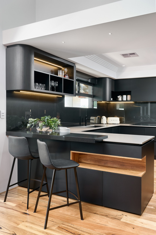 This is an example of a large contemporary u-shaped open plan kitchen in Perth with an undermount sink, flat-panel cabinets, black cabinets, quartz benchtops, black splashback, glass sheet splashback, black appliances, light hardwood floors, with island, brown floor, grey benchtop and recessed.