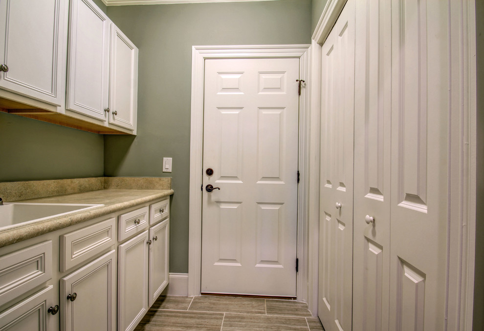 Photo of a large beach style galley dedicated laundry room in Wilmington with a drop-in sink, recessed-panel cabinets, white cabinets, laminate benchtops, ceramic floors, a concealed washer and dryer and grey walls.