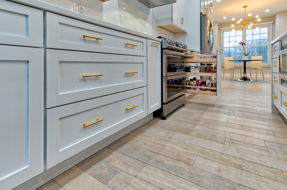 Inspiration for a mid-sized contemporary galley light wood floor and beige floor eat-in kitchen remodel in Philadelphia with a farmhouse sink, shaker cabinets, white cabinets, quartz countertops, white backsplash, glass tile backsplash, stainless steel appliances, an island and white countertops