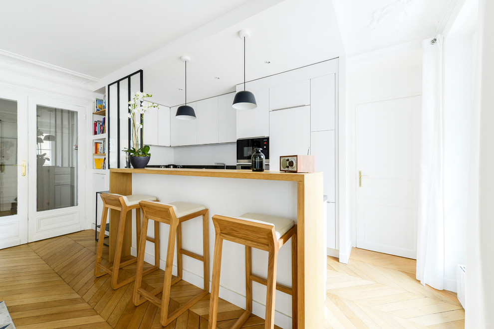 This is an example of a small contemporary u-shaped open plan kitchen in Paris with flat-panel cabinets, white cabinets, wood benchtops, white splashback, stainless steel appliances, light hardwood floors, a peninsula, brown floor, brown benchtop and an undermount sink.