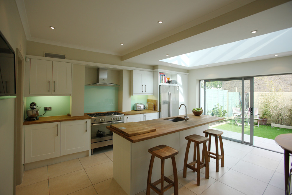 Photo of a mid-sized contemporary single-wall eat-in kitchen in London with an undermount sink, shaker cabinets, white cabinets, wood benchtops, green splashback, glass sheet splashback, stainless steel appliances, ceramic floors, with island, beige floor and brown benchtop.
