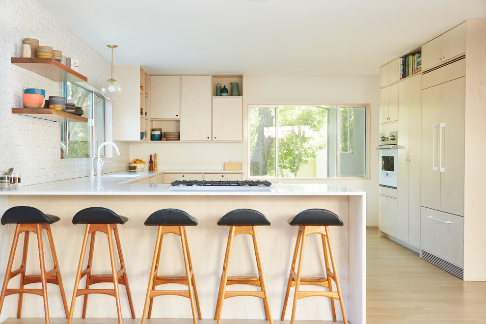 Design ideas for a midcentury u-shaped kitchen in Los Angeles with flat-panel cabinets, light wood cabinets, white splashback, ceramic splashback, panelled appliances, an integrated sink, light hardwood floors, a peninsula and beige floor.