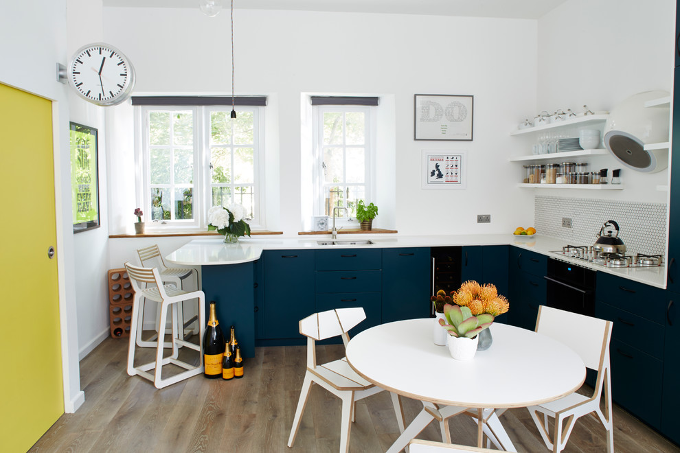 Photo of a contemporary l-shaped eat-in kitchen in London with an undermount sink, flat-panel cabinets, blue cabinets, quartz benchtops, white splashback, mosaic tile splashback, black appliances, medium hardwood floors and a peninsula.