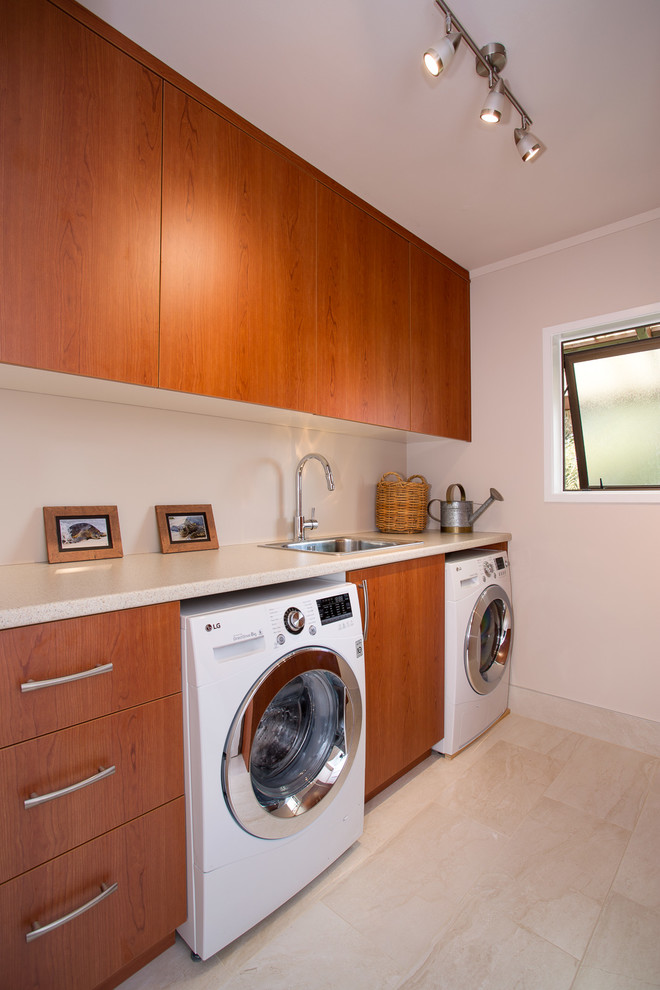 Photo of a large contemporary galley dedicated laundry room in Auckland with a drop-in sink, flat-panel cabinets, medium wood cabinets, beige walls, porcelain floors, a side-by-side washer and dryer and laminate benchtops.
