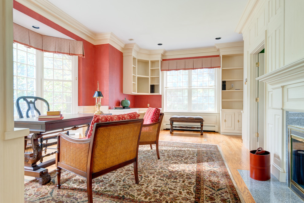 This is an example of a large home office in Boston with a library, red walls, medium hardwood floors, a standard fireplace, a wood fireplace surround and a freestanding desk.