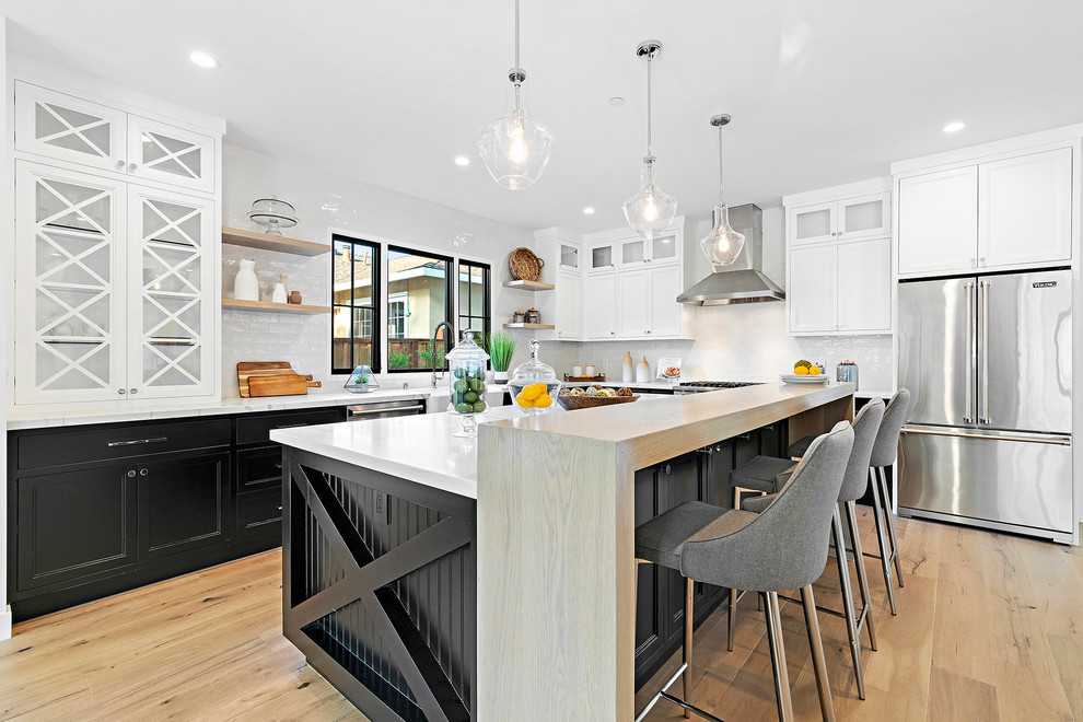 Photo of a large transitional l-shaped open plan kitchen in San Francisco with a farmhouse sink, recessed-panel cabinets, quartzite benchtops, white splashback, stone tile splashback, stainless steel appliances, with island, white benchtop, white cabinets, medium hardwood floors and brown floor.
