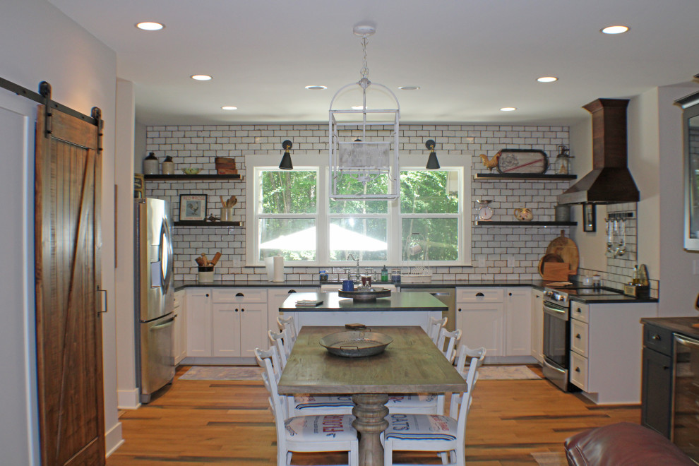 Inspiration for a mid-sized farmhouse u-shaped medium tone wood floor and brown floor eat-in kitchen remodel in Nashville with a farmhouse sink, flat-panel cabinets, white cabinets, granite countertops, white backsplash, subway tile backsplash, stainless steel appliances, an island and black countertops