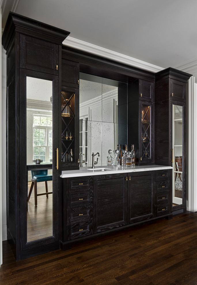 Photo of a mid-sized traditional galley wet bar in Detroit with an undermount sink, beaded inset cabinets, dark wood cabinets, marble benchtops, dark hardwood floors, mirror splashback, brown floor and white benchtop.